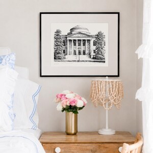 Op de afbeelding: Een zwart-wit architectuurprint in een zwarte lijst hangt boven een houten nachtkastje. Een gouden vaas bevat roze en witte rozen. Een witte lamp met een kralenkap staat op het nachtkastje. De sc&egrave;ne is in een slaapkamer.