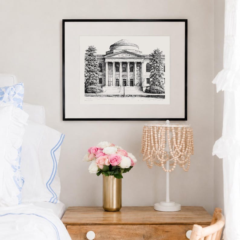 Op de afbeelding: Een zwart-wit architectuurprint in een zwarte lijst hangt boven een houten nachtkastje. Een gouden vaas bevat roze en witte rozen. Een witte lamp met een kralenkap staat op het nachtkastje. De sc&egrave;ne is in een slaapkamer.