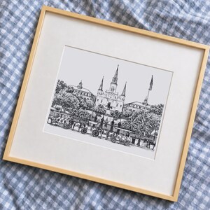 Könnte beinhalten: Eine gerahmte schwarz-weiße Tuschezeichnung der St. Louis Kathedrale in New Orleans. Die Zeichnung zeigt die Kathedrale mit ihren drei Türmen, eine Statue im Vordergrund, Pferdekutschen, Palmen und eine amerikanische Flagge.