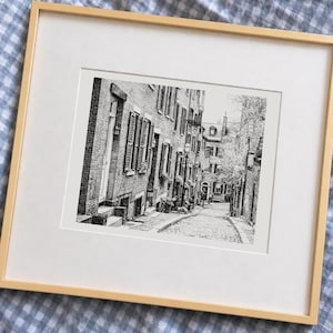 May include: A framed black and white ink drawing of a narrow cobblestone street lined with brick buildings, featuring shuttered windows, a hanging American flag, and a street lamp, presented in a light-colored wooden frame with a white mat.