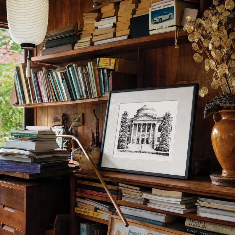 Op de afbeelding: Een ingelijste zwart-wit architecturale prent van een gebouw met zuilen en een koepel, tentoongesteld op een houten plank. De plank maakt deel uit van een ingebouwde boekenkast vol boeken. Een decoratieve vaas en een papieren lantaarn zijn ook zichtbaar.