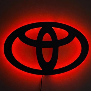 Könnte beinhalten: Ein schwarzes Toyota-Logo mit roter LED-Hintergrundbeleuchtung. Das Logo wird mit einem leuchtend roten Schein beleuchtet.