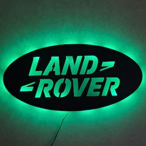 Puede incluir: Un letrero ovalado negro con las palabras "LAND-ROVER" en letras verdes. El letrero está iluminado con una luz verde.