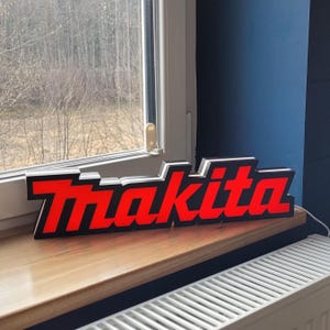 Könnte beinhalten: Ein beleuchtetes Schild mit dem Wort "Makita" in roten Buchstaben auf schwarzem Hintergrund.
