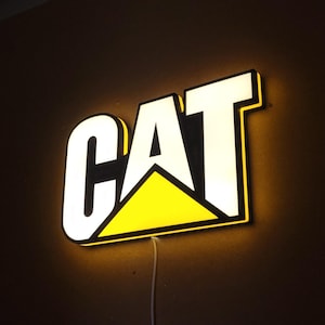 Könnte beinhalten: Beleuchtetes Schild mit dem Wort "CAT" in fetten, weißen Buchstaben und einem gelben Dreieckslogo. Das Schild ist schwarz umrandet und hat einen warmen, gelben Schein. Ein weißes Netzkabel erstreckt sich von unten.