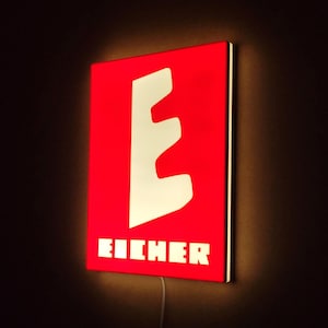 Op de afbeelding: Een rood rechthoekig bord met een witte letter 'E' en het woord 'EICHER' in witte blokletters. Het bord is verlicht, met een witte rand en een netsnoer aan de onderkant. Het geheel is bevestigd aan een donkere muur.
