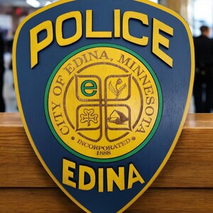 May include: A blue and yellow police badge with the words "Police" in yellow letters and "City of Edina, Minnesota" in a circle around a crest. The crest has a green shamrock, a building, and a flower. The words "Incorporated 1888" are below the crest. The words "Edina" are in yellow letters at the bottom of the badge.