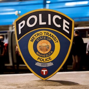 May include: A blue and yellow shield-shaped sign with the words "POLICE" in white letters at the top. The sign also says "METRO TRANSIT STATE OF MINNESOTA POLICE" and has the year "1993" on a white ribbon. A white "T" is in the center of the sign.