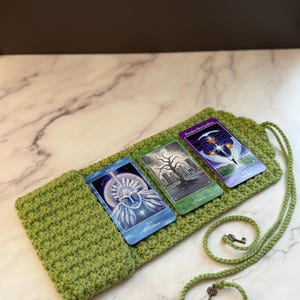 May include: A light green crocheted tarot card pouch with a long drawstring featuring two small metal key charms. Three tarot cards are placed on top of the pouch, displaying illustrations of a face, a tree, and a celestial figure.