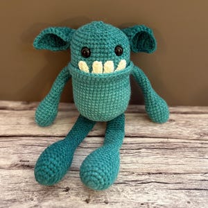 May include: A teal crocheted monster doll with large ears, black eyes, and a wide, toothy grin. The handmade plush toy has long arms and legs, and is sitting on a wooden surface. The doll is a whimsical, handmade item.