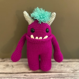 May include: A handmade magenta crochet monster toy with black button eyes, white teeth, and gray horns. The monster has a tuft of teal yarn on its head and outstretched arms. The toy is on a wooden surface.