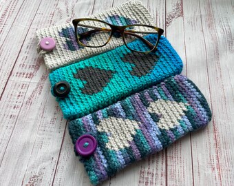 May include: Three handmade crochet eyeglass cases in shades of blue, teal, gray, and purple. Each case features a unique pattern and a button closure. A pair of eyeglasses rests on top of the cases.