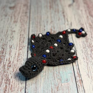 May include: A dark gray crocheted stingray toy featuring a flat, diamond-shaped body and a long, thin tail. The surface is decorated with small, round red, white, and blue beads. The stingray has a single visible eye with a blue iris and black pupil.