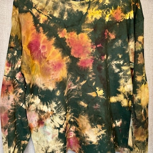 May include: A long-sleeved crew neck shirt featuring a tie-dye pattern in shades of dark forest green, golden yellow, and muted pink. The garment has a ribbed collar and long sleeves, with a swirling, abstract colour distribution across the fabric.