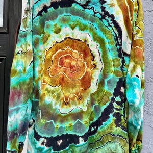 Op de afbeelding: Een lange mouwen tie-dye shirt met een spiraalpatroon. Het shirt heeft een mix van kleuren, waaronder teal, bruin, zwart en groen. Het ontwerp is gecentreerd en straalt naar buiten. Het shirt hangt aan een zwarte hanger.