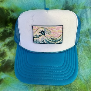 Könnte beinhalten: Blau-weiße Trucker-Cap mit weißer Front, blauem Mesh-Rücken und blauem Schirm. Auf der Front befindet sich ein rechteckiger Aufnäher mit einem gestickten Wellenmotiv in Blau-, Rosa- und Gelbtönen.
