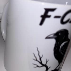 Op de afbeelding: Witte keramische mok met een zwarte ravenillustratie en de tekst "F-C". De raaf zit op een kale tak. De mok heeft een gebogen oor en is geschikt voor warme of koude dranken.