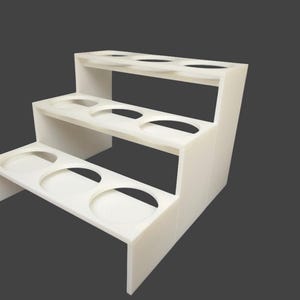 Op de afbeelding: Een witte, drielaagse displaystandaard met cirkelvormige uitsparingen op elk niveau. De standaard heeft een trapsgewijs ontwerp, waarbij elk niveau iets kleiner is dan het vorige. De standaard is gemaakt van een solide, ondoorzichtig materiaal.