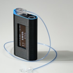 Könnte beinhalten: Ein schwarzes rechteckiges Gerät mit blauem Rand und einem kleinen Bildschirm, der Daten anzeigt. Auf der Oberseite befindet sich eine transparente Kunststoffkappe, die durch einen dünnen blauen Schlauch mit einem transparenten Kunststoffteil verbunden ist. Das Gerät befindet sich auf einer weißen Oberfläche.