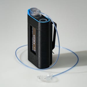 Może przedstawiać: Czarne urządzenie medyczne z niebieskim akcentem, przezroczystą rurką i małym ekranem wyświetlającym cyfry. Urządzenie ma klips z tyłu i przezroczystą nasadkę. Rurka jest połączona z przezroczystym, owalnym elementem.