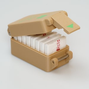 Könnte beinhalten: Eine rechteckige Box in Tan-Farbe mit einem Scharnierdeckel und einem Metallverschluss. Der Deckel ist geöffnet und zeigt einen Stapel weißer rechteckiger Tabletten im Inneren. Die Box hat einen grünen Pfeil auf der Seite.