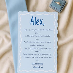 以下が含まれることがあります： 水色のネクタイと「Alex」という名前とメッセージが書かれたカード。カードにはスカラップの縁と水色の縁があります。青い箱に入った指輪も写っています。