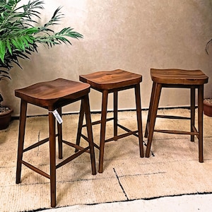 May include: Three wooden bar stools with a dark brown finish. Each stool has a contoured seat and four angled legs connected by a footrest. The stools are arranged on a textured rug, with plants in the background.