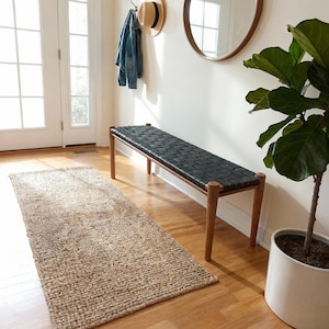 May include: A wooden bench with a woven black seat is in a bright hallway. The bench has four wooden legs and a rectangular shape. A natural fibre rug lies on the wooden floor. A denim jacket and a straw hat hang on the wall.