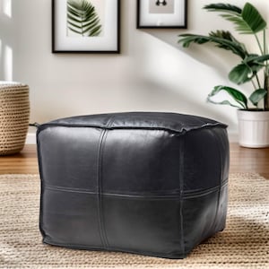May include: Black leather ottoman with a square shape and stitched panels. The ottoman is sitting on a rug.