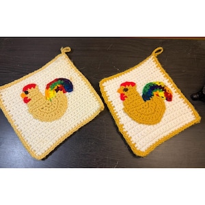 May include: Two square crochet potholders featuring an off-white base with a yellow border. Each potholder displays a centred, textured applique of a rooster with a tan body, a red comb, and a vibrant, multi-coloured tail in rainbow hues.
