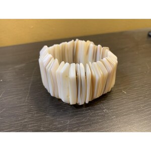 May include: A wide, ivory-colored bracelet composed of rectangular, shell-like pieces. The bracelet has a natural, organic appearance, with variations in color from cream to light brown. The pieces are strung together to form a flexible, chunky accessory.