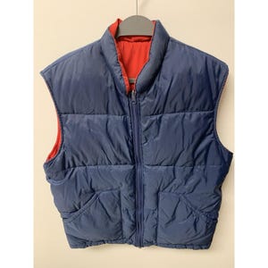 May include: A reversible puffer vest in navy blue with a red interior. The vest has a full zipper closure and two front pockets. The vest is hanging on a gray hanger.