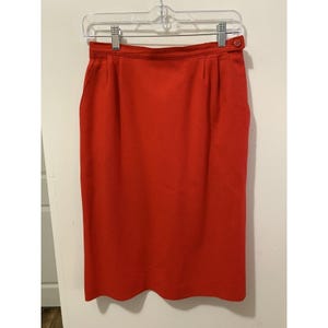 May include: A solid red midi-length skirt featuring a high waistband with a single button closure, subtle front pleats, and a straight silhouette. The skirt is suspended from a clear plastic hanger with two metal clips at the top waistband.