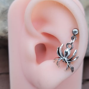 Könnte beinhalten: Ein silberfarbener Spinnen-Ohrhänger mit einem kleinen, klaren Schmuckstein als Stecker. Der Spinnenanhänger hängt an einem Biegering, der am Stecker befestigt ist, welcher in einem hautfarbenen Silikon-Ohr-Modell steckt. Die Spinne hat acht Beine.