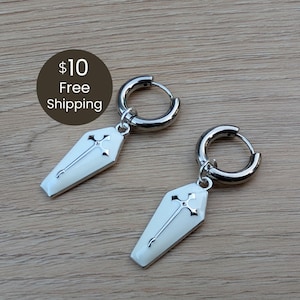 May include: A pair of silver-toned hoop earrings featuring dangling coffin-shaped charms. Each charm is finished with a smooth white enamel surface and a centered silver-toned cross emblem. The earrings have a polished metallic finish throughout.