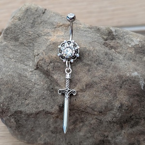 May include: A silver-colored belly button ring with a floral design and a dangling sword charm. The top ball and center of the floral design are adorned with clear crystals. The sword charm has intricate details.