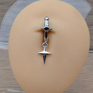 May include: A silver-colored belly button ring featuring a dagger design. The ring has a straight barbell with a small black gem and a dangling dagger pendant with a star-shaped hilt.