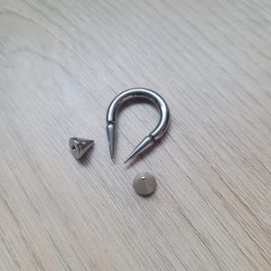 May include: A silver-colored, U-shaped body jewelry piece with two pointed ends, along with two separate, cone-shaped pieces. The jewelry is displayed on a light-colored wooden surface.