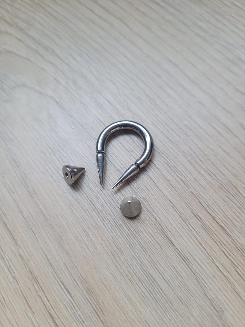 May include: A silver-colored, U-shaped body jewelry piece with two pointed ends, along with two separate, cone-shaped pieces. The jewelry is displayed on a light-colored wooden surface.