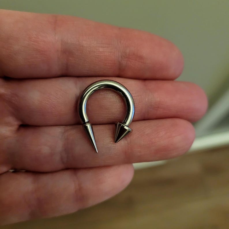 May include: A silver-colored horseshoe-shaped piercing with two pointed ends. The piercing is made of a shiny metal and is designed to be worn in a body piercing. The ends are cone-shaped.