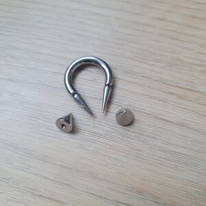 May include: A collection of silver-colored body jewelry, including a horseshoe-shaped barbell with pointed ends, a cone-shaped stud, and a small, round stud. The pieces are arranged on a light wood-grain surface.