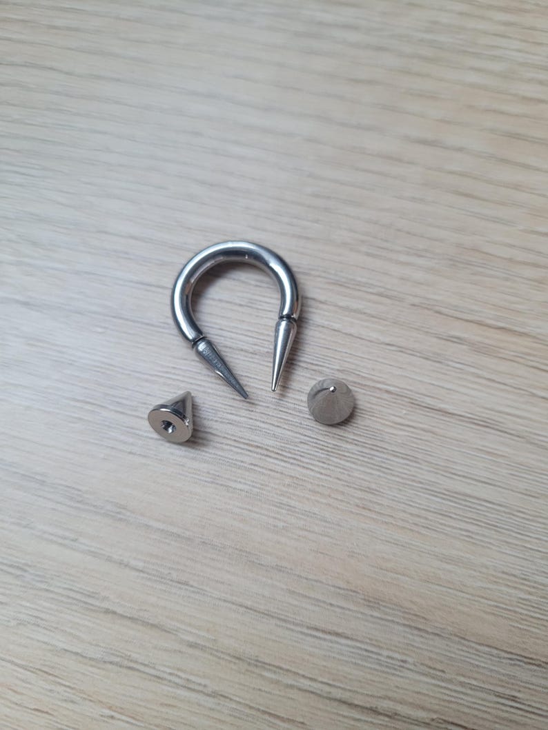 May include: A collection of silver-colored body jewelry, including a horseshoe-shaped barbell with pointed ends, a cone-shaped stud, and a small, round stud. The pieces are arranged on a light wood-grain surface.