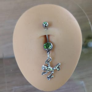 May include: A silver belly button ring with a frog charm. The ring has a green gem at the bottom and a smaller green gem at the top. The frog charm is detailed and adds a unique touch to the jewellery.