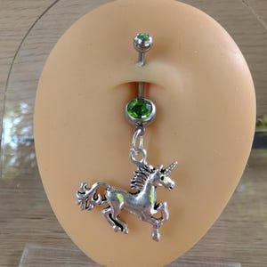 May include: A silver-colored belly button ring with a dangling unicorn charm. The ring features two green gemstone accents. The unicorn charm is detailed with a mane, tail, and horn.