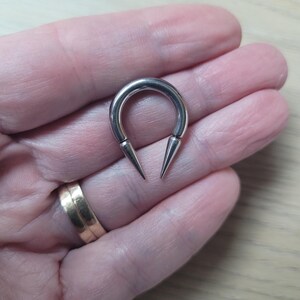 May include: A silver-colored, horseshoe-shaped ear gauge with two sharp, conical spikes extending from the ends. The gauge is held in a hand, and the design is simple and modern.