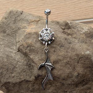 May include: A silver-colored belly button ring with a floral design and a dangling bat wing charm. The ring features a large, clear crystal center stone and smaller iridescent crystals around the floral design.