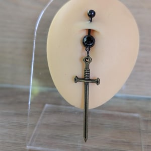 May include: A bronze-colored belly button ring featuring a sword pendant. The ring has a black gem and a decorative top ball. The sword pendant is detailed with a hilt and blade.