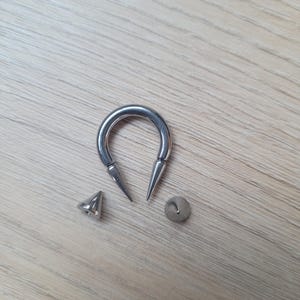 May include: A silver-colored, horseshoe-shaped ear gauge with pointed ends, alongside a matching spike and a small, round backing. The jewelry is designed for stretched ear piercings.