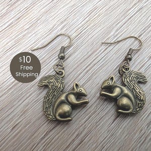 May include: A pair of antique bronze-colored squirrel earrings. Each earring features a detailed squirrel charm with a bushy tail and a hook closure. The earrings are displayed on a light wood-grain surface.
