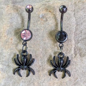 Könnte beinhalten: Zwei schwarze Bauchnabelpiercings mit gebogenem Steg-Design. Eines hat einen rosa Edelstein und das andere einen schwarzen Edelstein. Jeder Ring hat einen hängenden schwarzen Spinnenanhänger, der einen einzigartigen, gotischen Flair verleiht.
